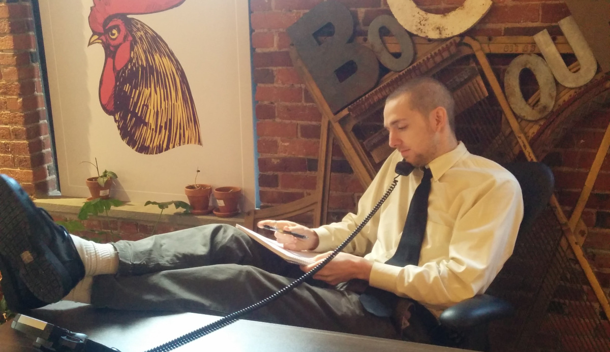 The author seated, feet on a desk and cradling a corded phone on his shoulder. In the background, the word "Bocoup" is spelled in iron lettering alongside a print-out of the Bocoup mascot