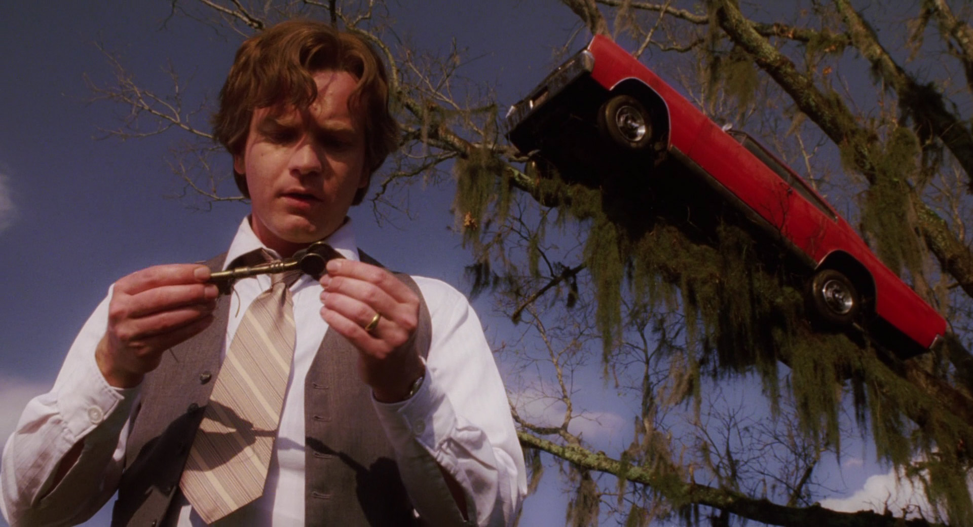 A still from the 2003 film "Big Fush" in which protagonist Edward Bloom inspects a key while behind him, a car hangs suspended in the branches of a tree.